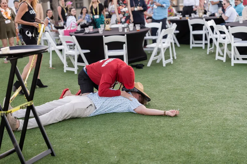 Two guests wrestle playfully on the lawn, one in red and one in light blue, surrounded by white chairs.
