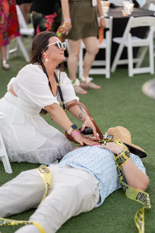 A woman in white stretches across another guest's legs during a playful moment on the grass.