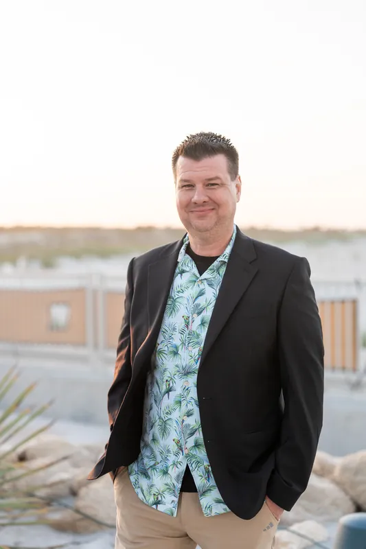 A man in a black blazer and floral shirt stands confidently with the waterfront visible behind him.