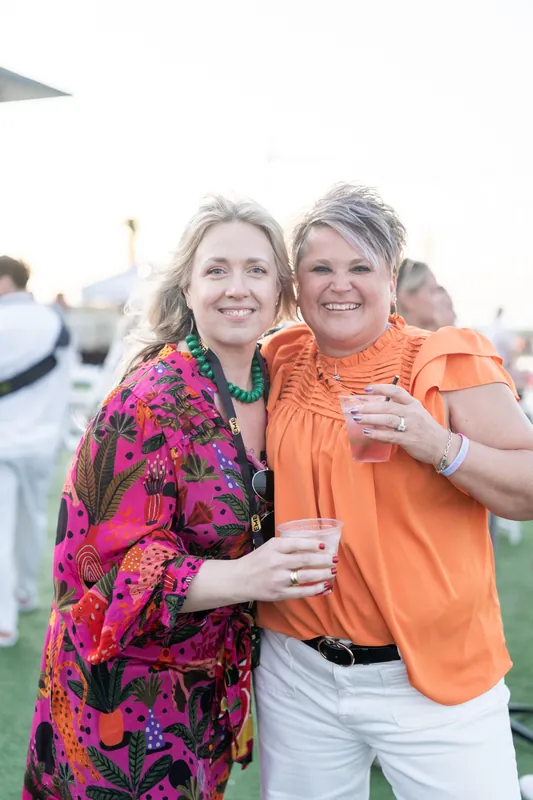 Two women in vibrant patterned outfits hold drinks at an outdoor beach event, smiling warmly toward the camera.