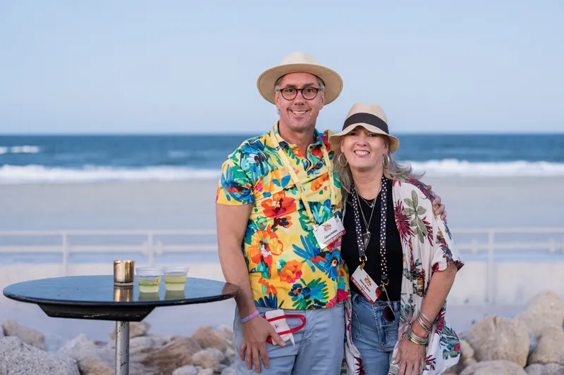 A man in a tropical floral shirt and woman in a black jacket pose together with the ocean behind them at the beach.