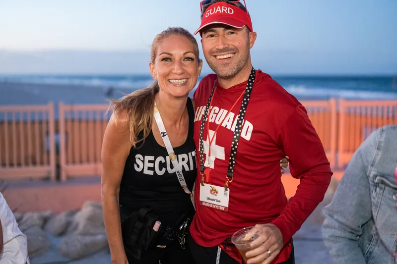 A woman in a black tank top and man in a red cap embrace while standing on a beachside deck at sunset.