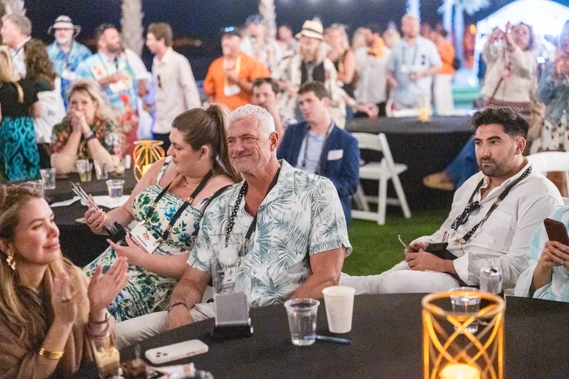 Guests seated at tables enjoy an evening reception under string lights and tropical décor on the lawn.
