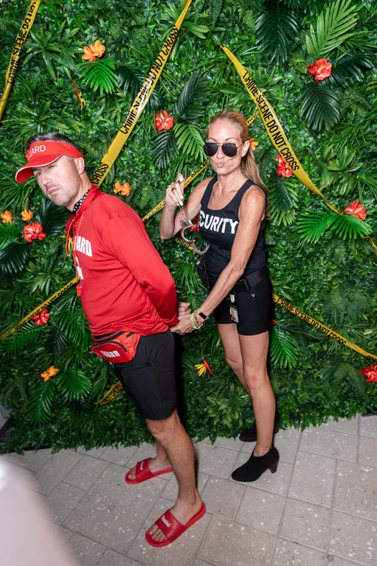A man in red and woman in black sunglasses pose playfully in front of a lush green floral backdrop.