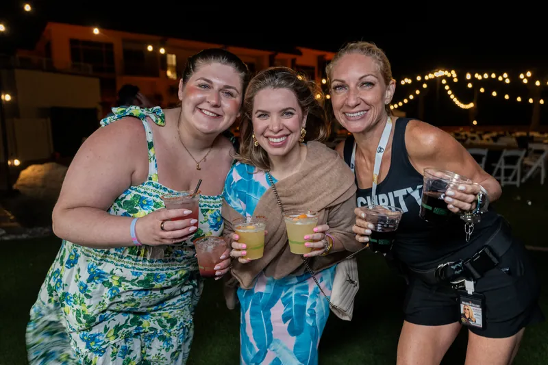 Three women in colorful summer outfits hold drinks and smile at an evening event at Embassy Suites St. Augustine Beach.