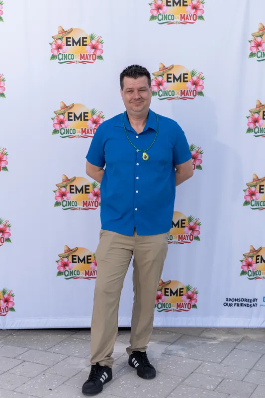 Man wearing a bright blue button-up shirt and tan pants stands with arms outstretched against an EME branded backdrop.