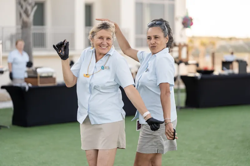 Two women in white shirts and dark gloves pose on a grass lawn with a coastal venue visible behind them.
