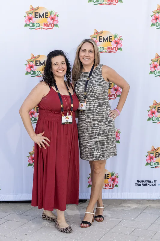 Woman in a burgundy dress and woman in a gray patterned dress stand together at the EME event photo backdrop.