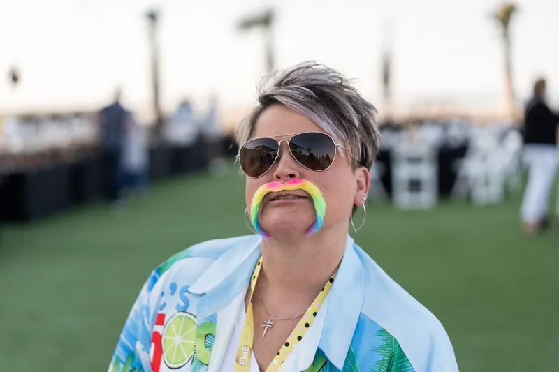 Guest wearing sunglasses and a colorful patterned shirt grins while holding a green prop mustache on a field.