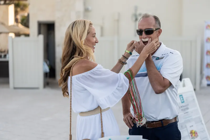 A man in sunglasses playfully adjusts a woman's white off-shoulder dress during an outdoor event at the beach resort.