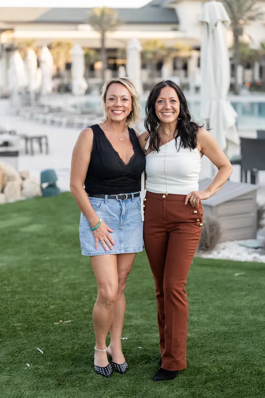 Two women stand arm-in-arm on the lawn, one in a black top and denim, the other in a white tank and rust-colored pants.
