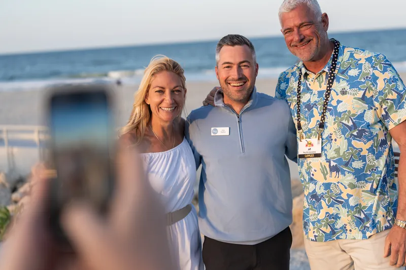 Three guests smile at the ocean backdrop, with the man in the center wearing a blue polo shirt and name badge.