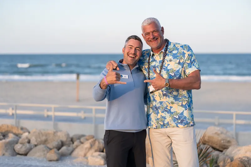 Two guests give thumbs up on the beach with waves and sky visible behind them, one in a blue shirt and the other in a floral