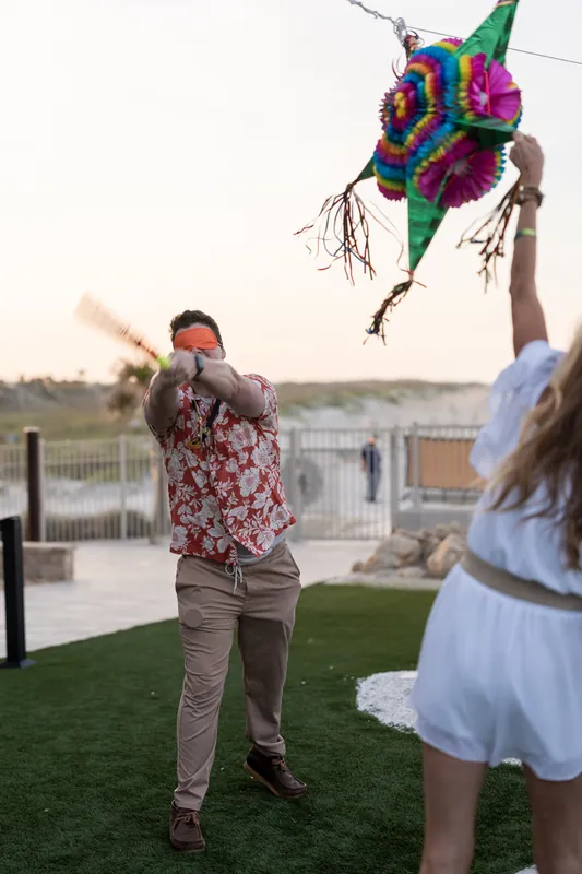 Man in floral shirt launches a colorful piñata skyward on the Embassy Suites beach terrace as a woman in white watches.