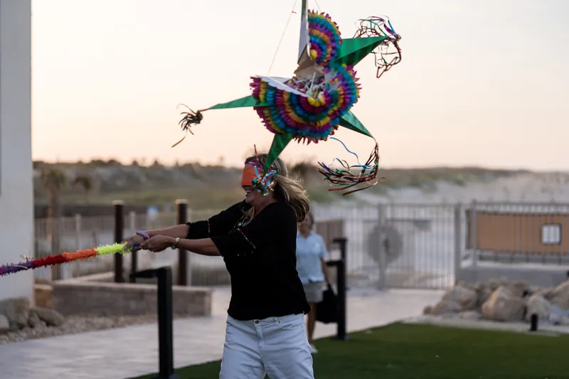 Woman in black jacket catches a vibrant piñata mid-air with the beach and waterfront buildings behind her.