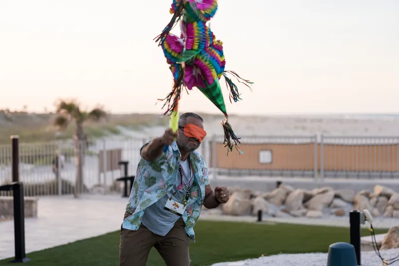 Man in light blue shirt swings at a dangling piñata during an outdoor celebration overlooking the St. Augustine Beach.