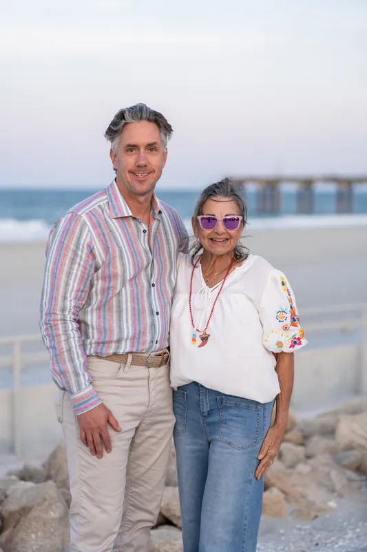 Couple stands together on the beach with the ocean and pier visible behind them at sunset.