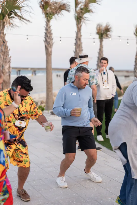 Guests in casual attire run and celebrate on the oceanfront terrace with palm trees framing the scene.