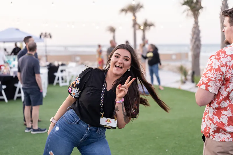 Woman in a black blazer and jeans laughs while making a peace sign at a beachside event.