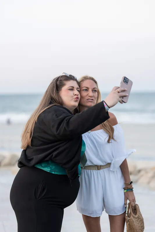 Two women take a selfie together on the beach with the ocean and sand behind them.