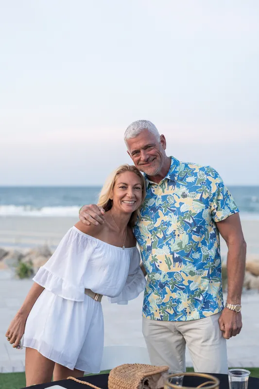 A couple poses arm-in-arm on the beach in casual summer clothing overlooking the water.