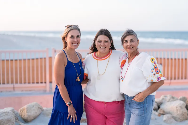 Three women stand together smiling for a photo on a beach deck with the ocean visible behind them.