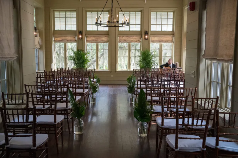 Wooden chiavari chairs line both sides of an aisle at Oyster Bay Yacht Club, with tall windows and green plants framing