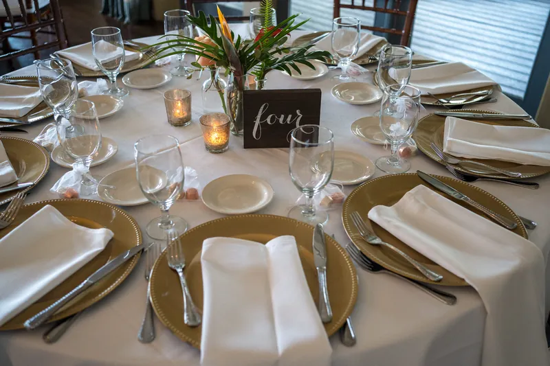 Round table set for dinner with gold chargers, white napkins, glassware, and a small chalkboard reading "four" at Oyster Bay