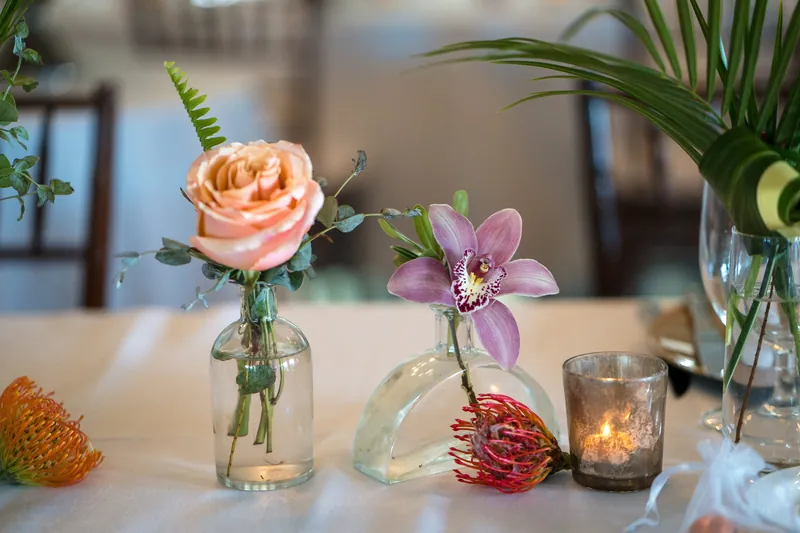 Multiple bud vases on a white table feature a peach rose, purple orchid, and deep red dahlia with candles and greenery.