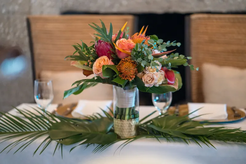Vibrant floral arrangement with orange dahlias, peach roses, and pink flowers in a clear vase on a white banquet table.