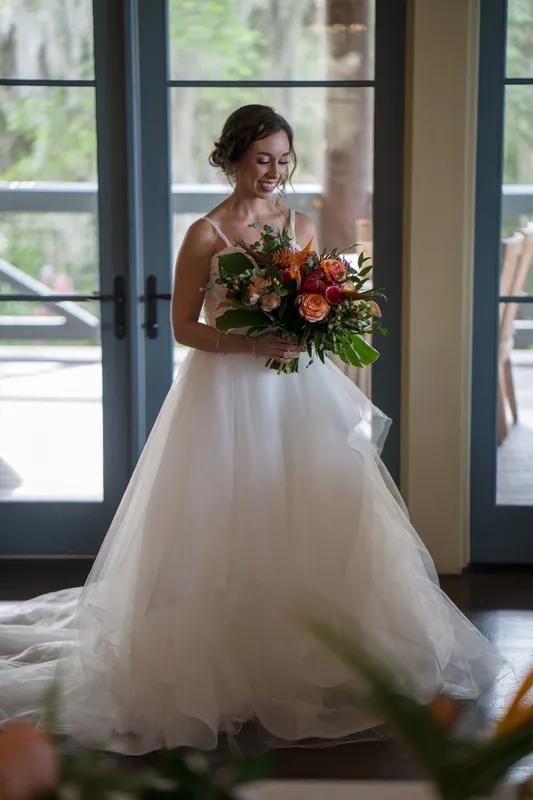 Emma stands in a flowing white gown holding her bouquet near tall windows overlooking the waterfront venue.