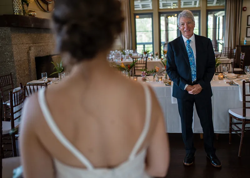 Kyle in a dark suit and blue tie smiles as Emma, seen from behind in her wedding dress, greets him.