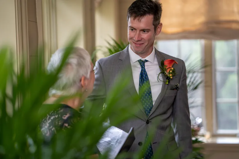 Kyle wears a gray suit with a floral boutonniere, framed by potted greenery in the yacht club's light-filled space.