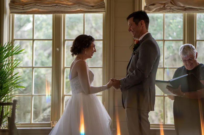 Emma in a white wedding dress holds Kyle's hands as they stand before tall windows during their first look moment.
