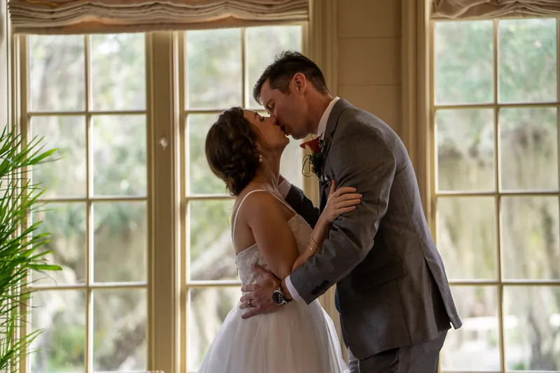 Emma and Kyle kiss at the altar with tall windows and greenery visible behind them at their yacht club ceremony.