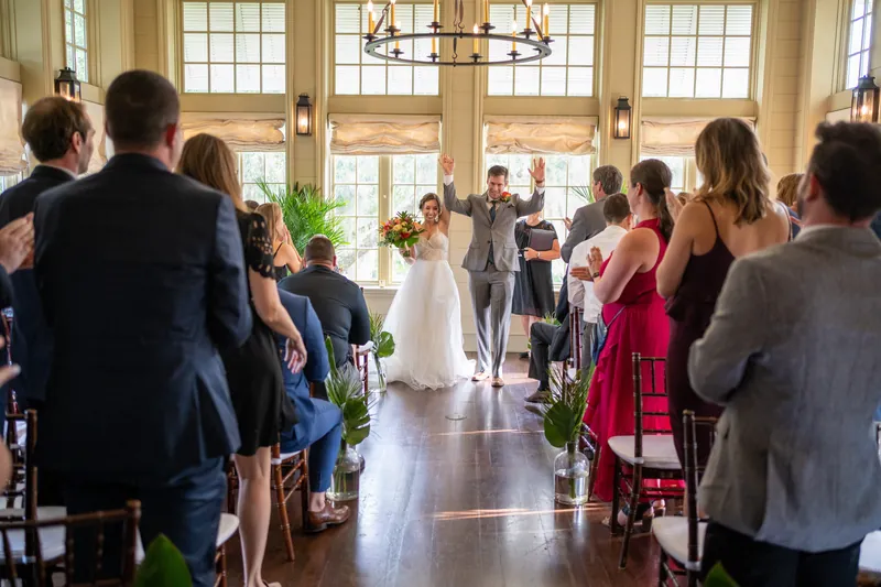 Emma in a white gown walks down the aisle at Oyster Bay Yacht Club as guests seated in blue chairs watch the ceremony unfold.