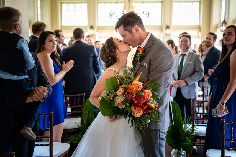 Kyle leans in to kiss Emma during their ceremony, as she holds an orange and coral bouquet and wedding guests stand nearby.