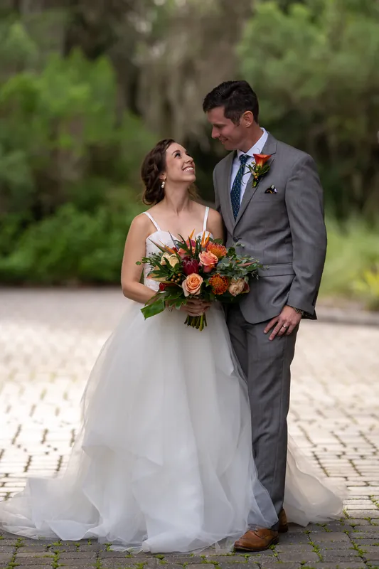 Emma looks up at Kyle while holding her orange and coral bouquet on a brick pathway lined with moss-draped trees.