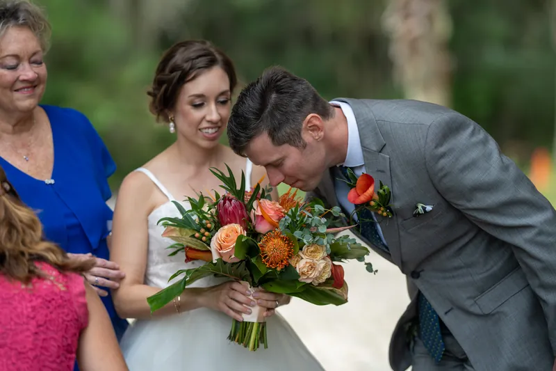 Kyle bends to kiss Emma's bouquet of red and orange roses while family members smile nearby at the yacht club.