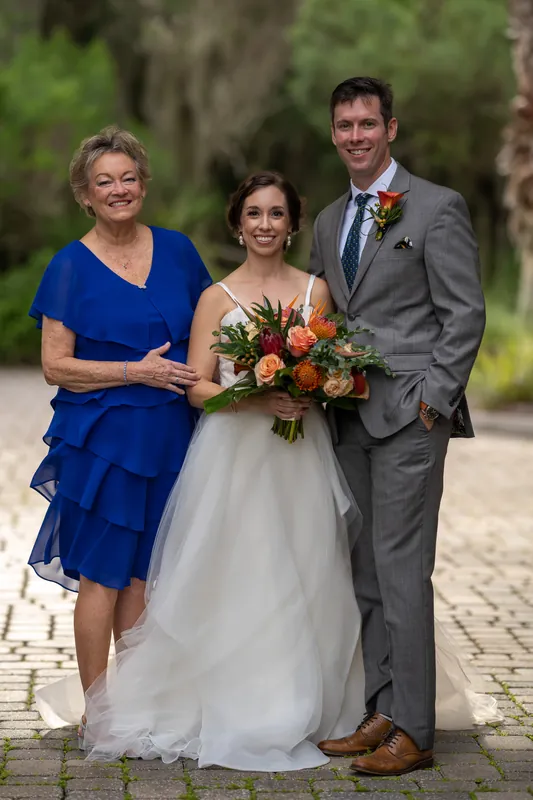 Emma holds her vibrant red and orange bouquet while posing with Kyle and a family member on a brick pathway.