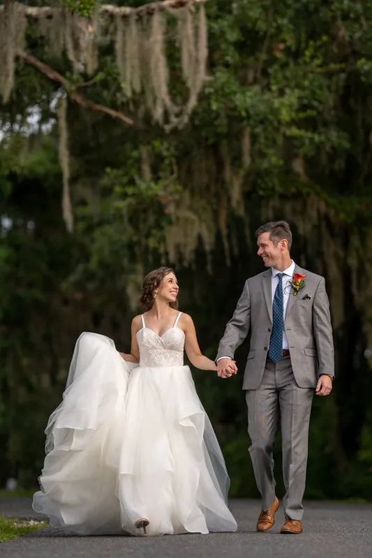 Emma and Kyle walk hand-in-hand down a tree-lined path draped with Spanish moss, Emma's white tulle gown flowing behind them.