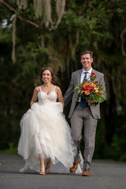 Kyle and Emma stroll together along a brick path beneath hanging Spanish moss, Emma's dress billowing as they hold hands.