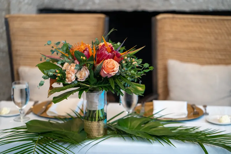 Centerpiece of coral, orange, and pink roses mixed with tropical greenery sits on a white linen table set with place settings