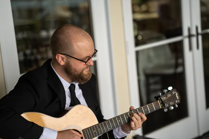Musician in black suit and glasses plays acoustic guitar indoors at the yacht club reception.
