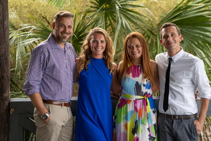 Four guests pose together outdoors, smiling at the camera with palm fronds and the yacht club visible behind them.