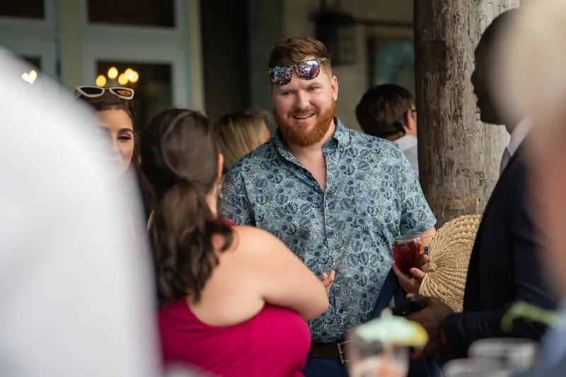 Kyle in a blue patterned shirt holds a drink while conversing with guests during the evening reception.