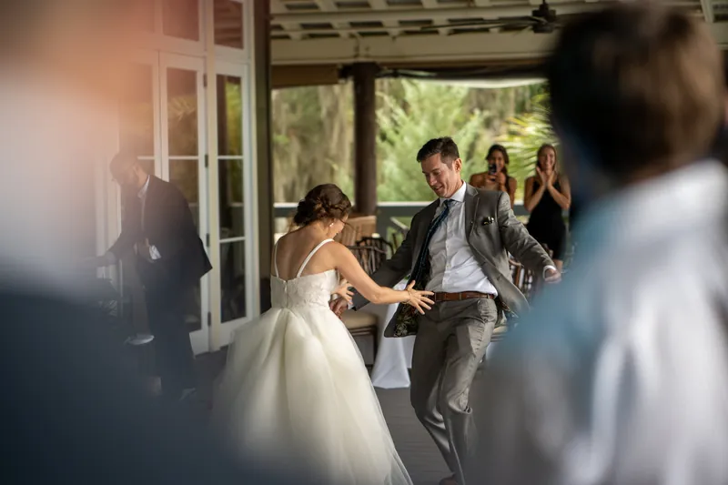 Kyle and Emma laugh while dancing together indoors as guests watch through glass doors and windows.
