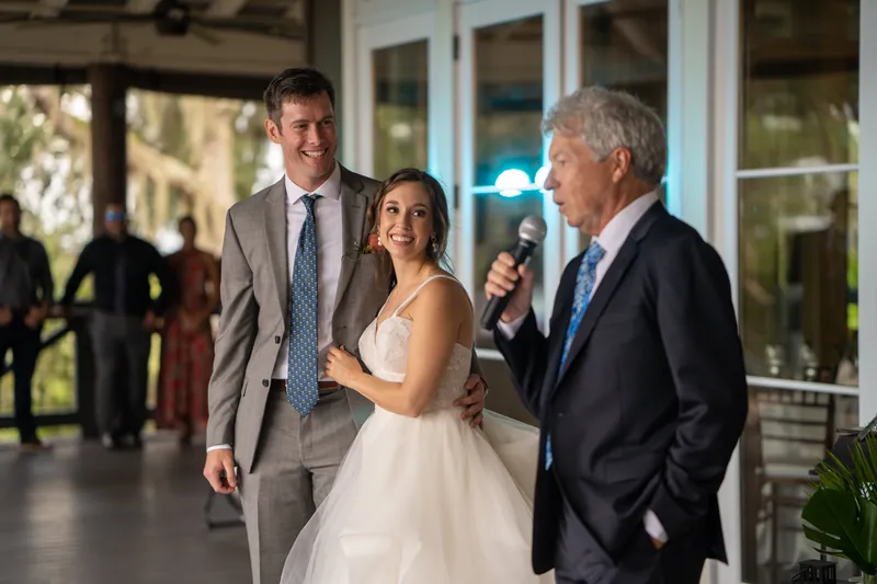 Emma stands between Kyle and a man holding a microphone, both smiling during toasts at the reception.