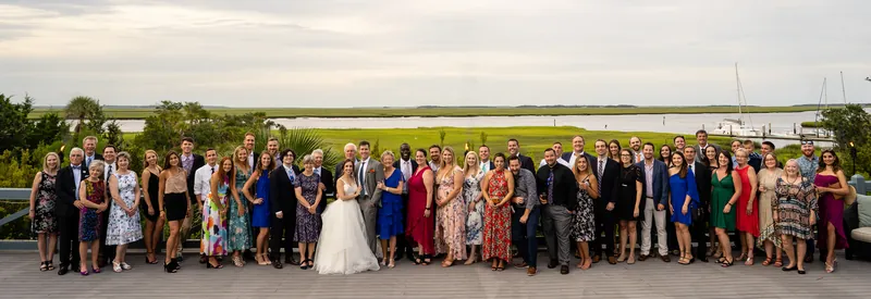 The full wedding party and guests gather outdoors for a group portrait with the water and sky behind them.