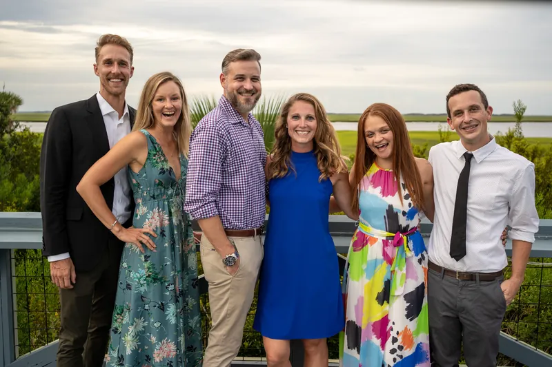 Six guests in mixed formal attire stand together by the water at Oyster Bay Yacht Club, with waterfront and marsh grasses